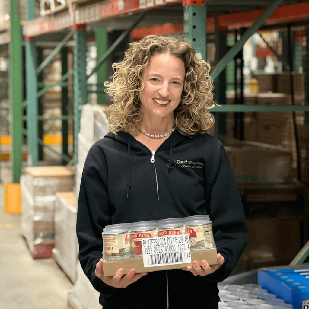 Heather Paquette, president of Good Shepherd Food Bank, in the Auburn Distribution Center, holding a case of cans in a black sweatshirt