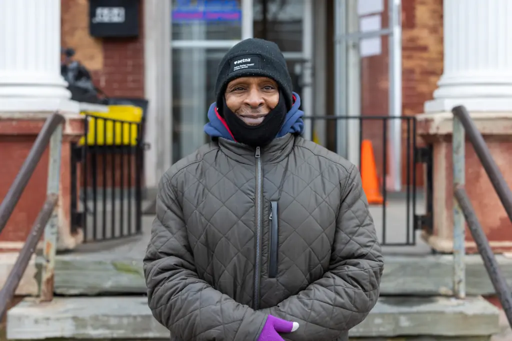 Raymond - Feeding America story - Man in front of door with puffy brown jacket on and purple gloves with a black winter hat.