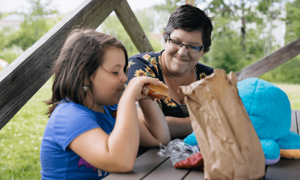 A woman smiling at a child eating a sandwich.