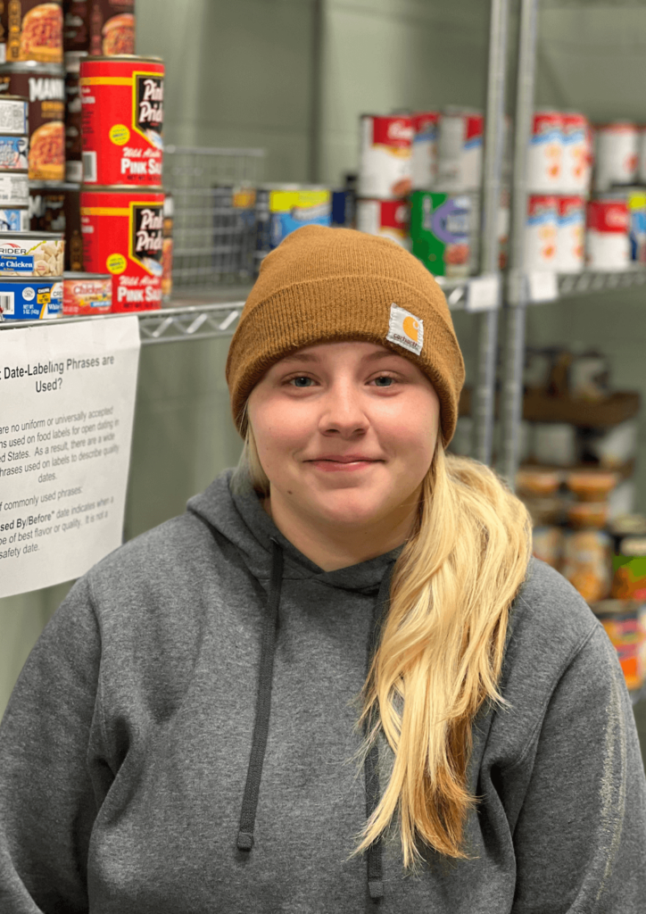 A woman with blond hair and a hat standing in front of food shelves.