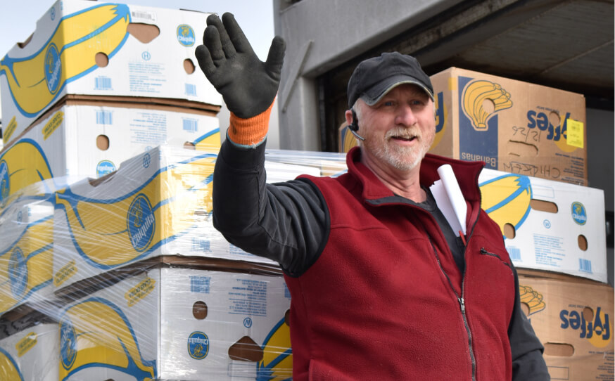 A man waving standing in front of a box of bananas